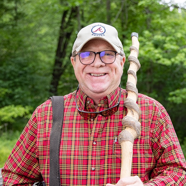 A smiling man wearing glasses, a tan baseball cap, and a red plaid shirt stands outdoors holding a large, twisted wooden walking stick, with green trees in the background.