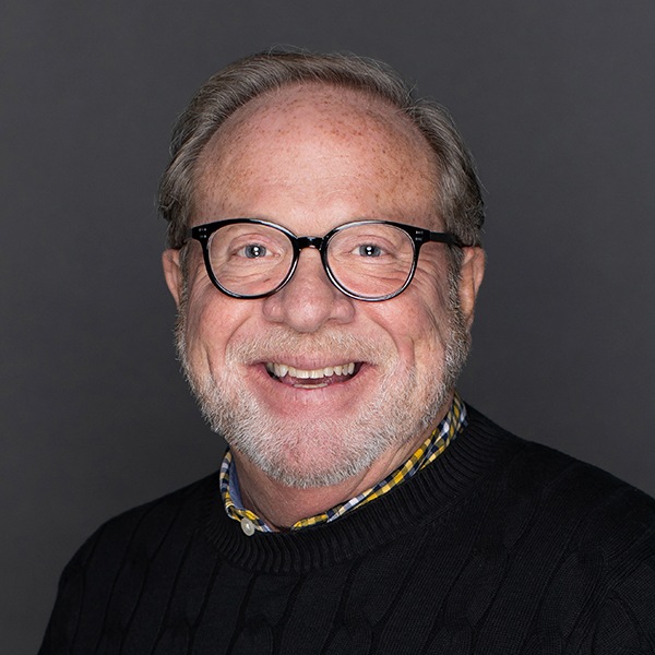 A smiling older man with light skin, gray hair, glasses, and a beard, wearing a black sweater over a checkered shirt, posed against a plain dark background.
