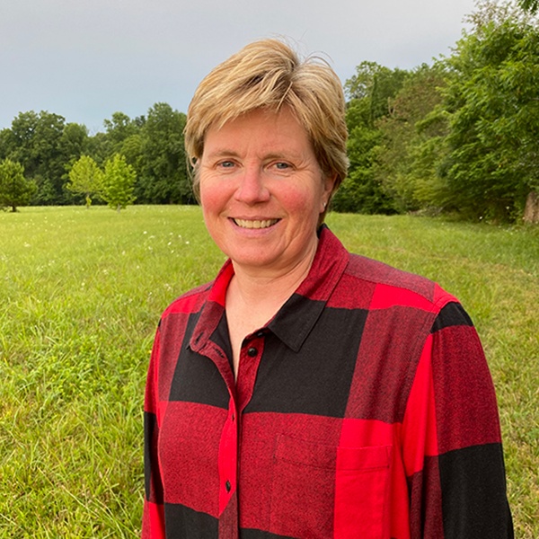 A woman with short blonde hair wearing a red and black plaid shirt stands smiling in a grassy field with trees in the background under a cloudy sky.
