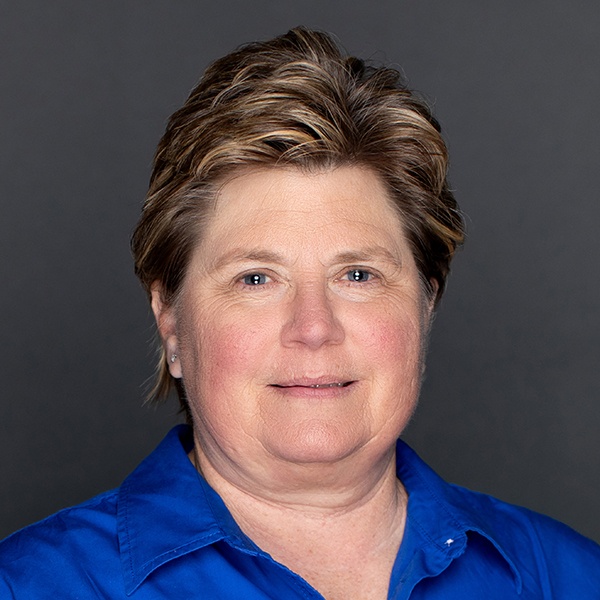 A middle-aged woman with short, light brown hair, blue eyes, and fair skin, wearing a bright blue collared shirt, smiles softly against a plain dark gray background.
