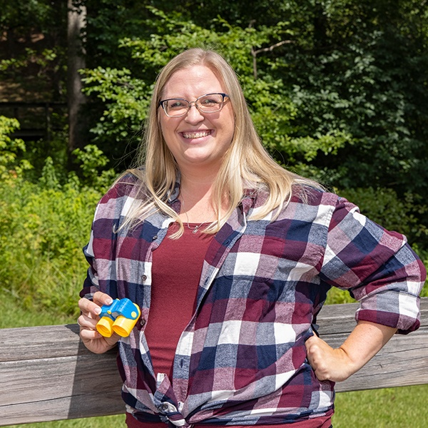 A smiling woman with long blonde hair and glasses stands outdoors, wearing a red and blue plaid shirt over a maroon top. She holds a pair of toy binoculars and stands in front of a wooden railing with greenery in the background.