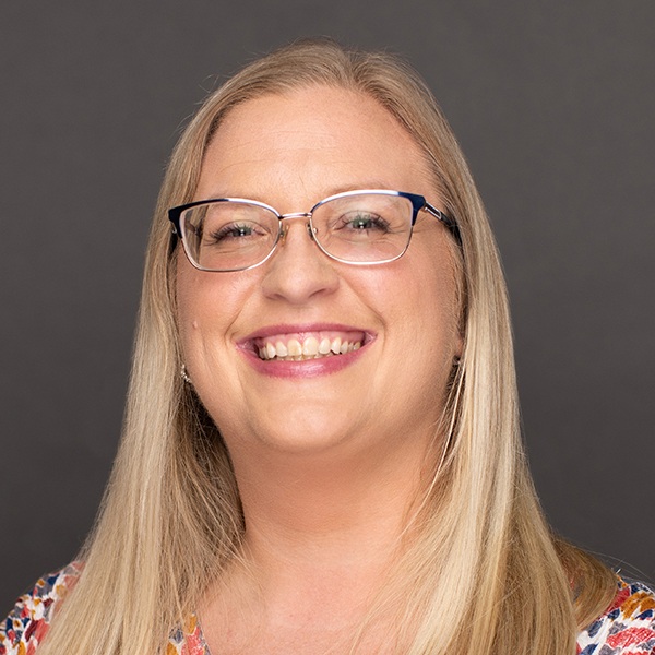 A woman with long blonde hair and glasses smiles at the camera. She is wearing a colorful patterned top, and the background is plain and dark gray.