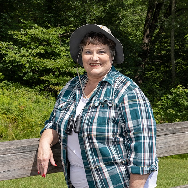 A smiling woman wearing a plaid shirt, hat, and binoculars around her neck leans on a wooden fence outdoors, with green trees and grass in the background.