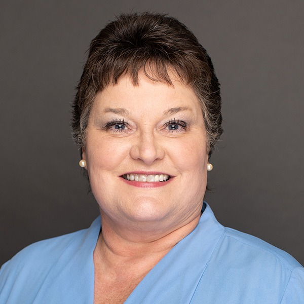 Smiling woman with short brown hair, wearing pearl earrings and a light blue collared blouse, posed against a plain gray background.