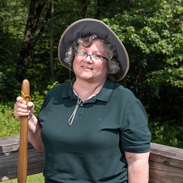 A smiling woman with gray hair wearing glasses, a wide-brimmed hat, and a dark green polo shirt stands outdoors on a wooden bridge holding a walking stick. Lush green trees are visible in the background.