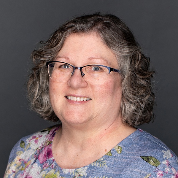 Smiling woman with short, curly gray hair and glasses, wearing a blue patterned top with floral designs, posed against a plain dark gray background.