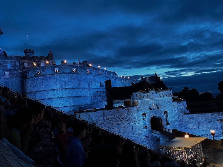 Edinburgh Castle at dusk, with the fortress walls lit up against a dark blue sky; people are seated in the foreground to watch an event near the castle gates.