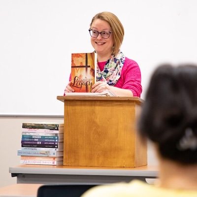A woman with glasses and a pink sweater stands at a podium, smiling and holding up a book. A stack of books is on the podium and another stack is on a nearby table. A person is seated in the foreground.
