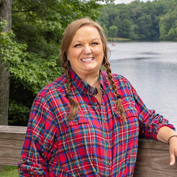 A woman with braided hair, wearing a red plaid shirt, stands smiling by a wooden railing with a lake and green trees in the background.