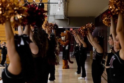 Cheerleaders in black uniforms holding pom-poms line a gymnasium, while a person in a wildcat mascot costume stands in the background, clapping and facing the group.