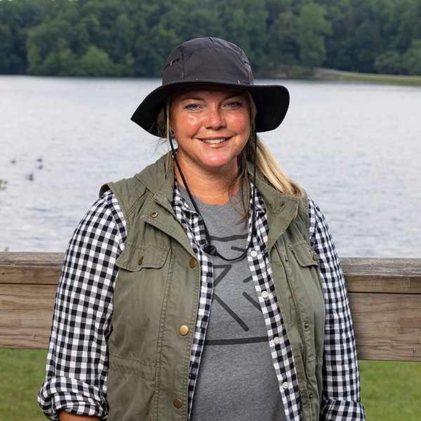 A smiling woman in an outdoor setting wears a black hat, green vest, and black-and-white checked shirt, standing in front of a wooden fence by a lake with trees in the background.