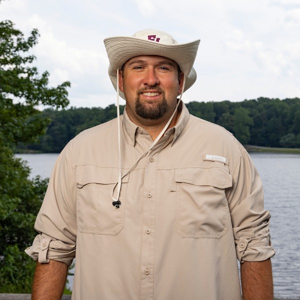 A man wearing a light-colored wide-brimmed hat and a beige button-up shirt stands outdoors in front of a lake with trees in the background, smiling at the camera.
