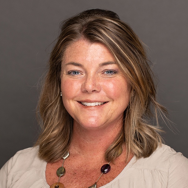 Smiling woman with shoulder-length light brown hair, wearing a cream blouse and a chunky beaded necklace, posed in front of a plain dark background.