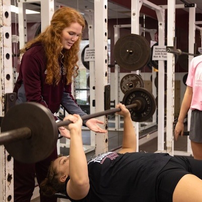 A coach with long red hair spots a person performing a bench press in a gym. Another person stands nearby. Weightlifting equipment surrounds them.