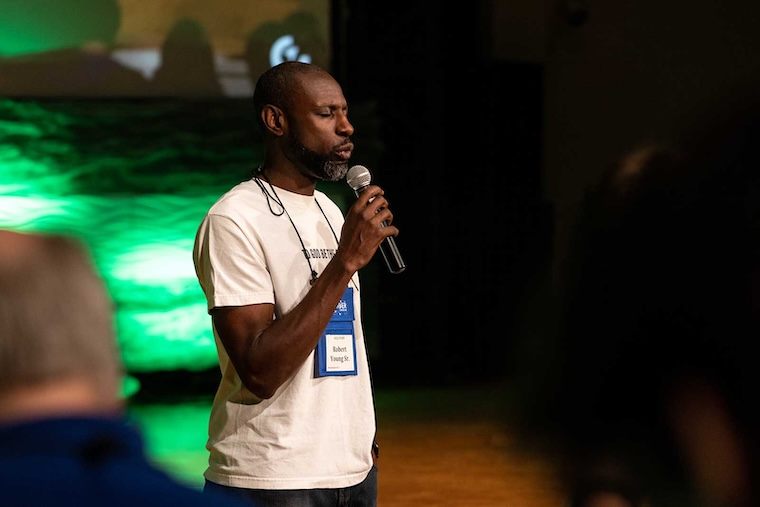 A man wearing a white t-shirt and conference badge speaks into a microphone on stage, with a green-lit background and audience members visible in the foreground.