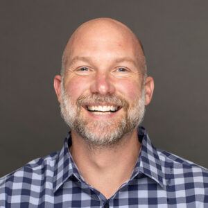 A smiling bald man with a short beard and mustache, wearing a blue and white checkered shirt, poses against a plain gray background.