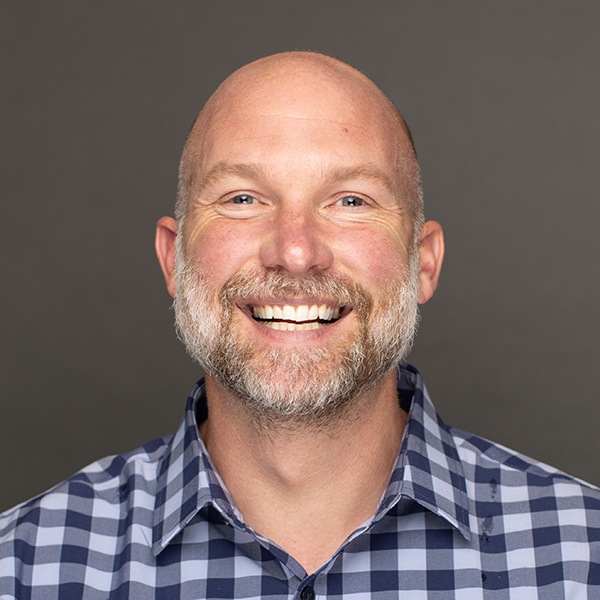 A smiling bald man with a short beard and mustache, wearing a blue and white checkered shirt, poses against a plain gray background.