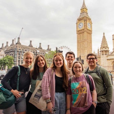 Six smiling young adults pose together in front of Big Ben in London on a cloudy day, with historic buildings and the London Eye visible in the background.