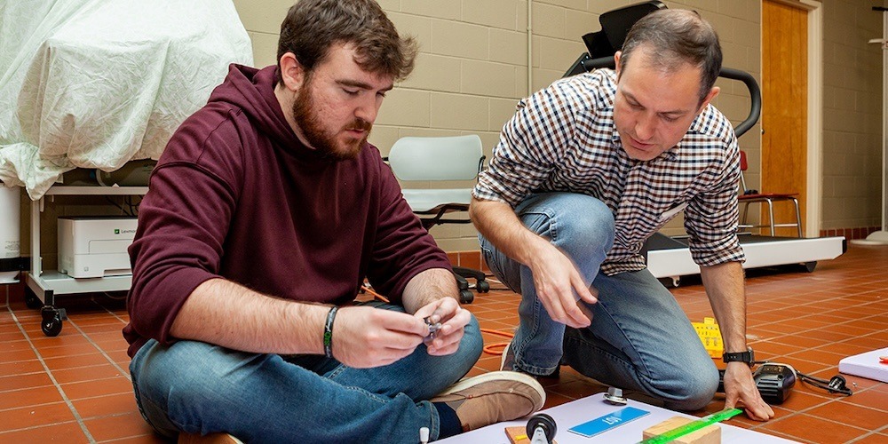 Two men kneel on the floor working on a project with papers, tools, and small objects. One wears a hoodie and jeans; the other wears a checkered shirt and khakis. They appear focused in a classroom or lab setting.