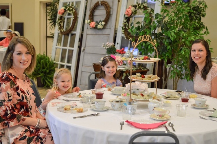Two young girls in floral dresses sit smiling at a round table set for tea, with tiered trays of treats and teacups. The background features vintage doors decorated with wreaths and flowers.