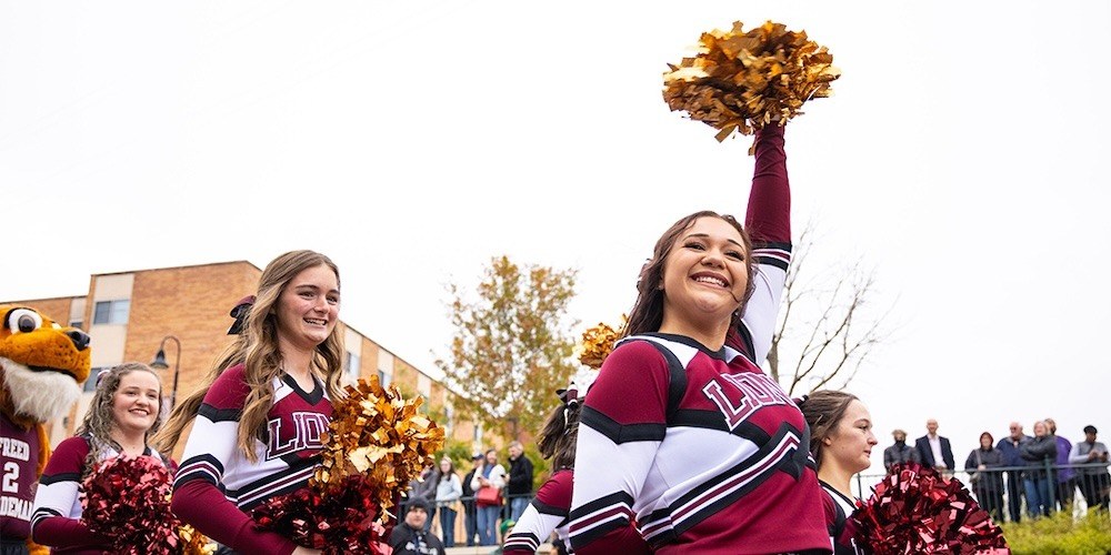 A group of cheerleaders in maroon and white uniforms, one raising gold pom-poms, smile during an outdoor event. People and a mascot are visible in the background near a brick building and trees.