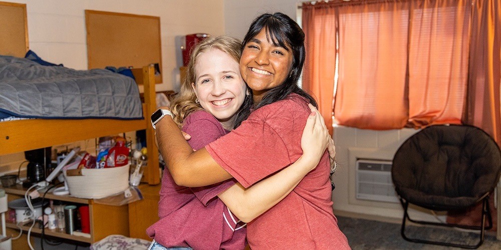 Two smiling young women hug each other in a dorm room with a bunk bed, shelves, and an orange curtain, both wearing casual shirts and appearing happy.
