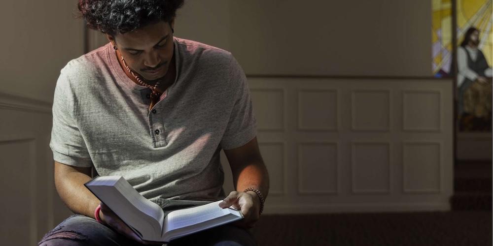 A man sits indoors reading a book in soft lighting. He appears focused, with a stained glass window featuring a religious figure visible in the background.
