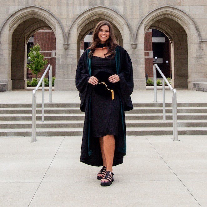 Dr. Olivia Gregory in academic regalia, standing in front of arched stone architecture on the FHU campus, holding her graduation cap and smiling.