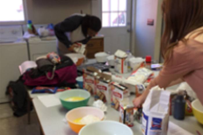 Two people are preparing ingredients in a kitchen with various baking supplies, bowls, and flour bags spread out on a table. The image is blurry.