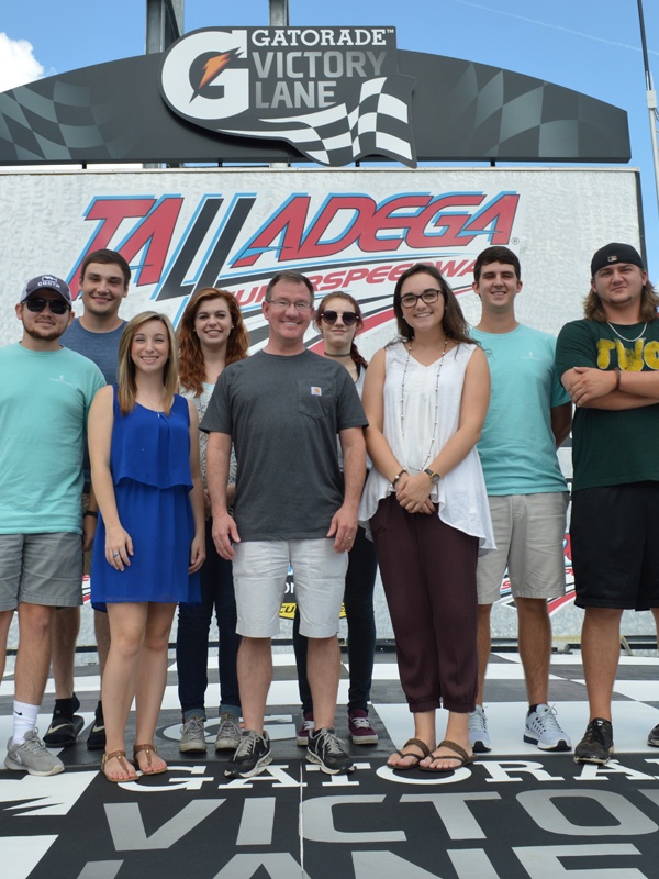 A group of nine people smile and pose together in front of a Gatorade Victory Lane sign at Talladega Superspeedway on a sunny day. The racetrack logo is visible behind them.