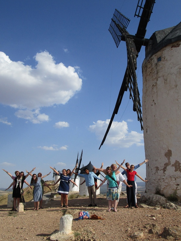 A group of people stand in front of old windmills on a sunny day, holding their arms outstretched. The sky is blue with scattered clouds, and the landscape appears dry and rocky.