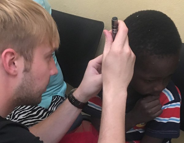 A man uses a small flashlight to examine a young boy’s ear while the boy sits quietly, looking down and wearing a striped shirt.