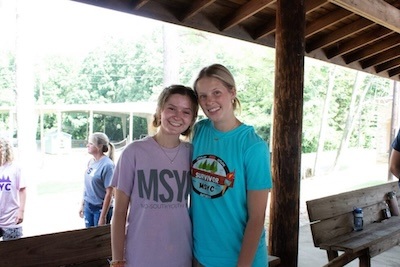Two young women stand smiling and hugging under a wooden pavilion. One wears a light purple MSYC shirt, and the other wears a blue Survivor camp shirt. Other people and trees are visible in the sunny background.