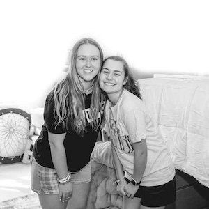 Two young women smiling and posing together in a dorm room. One is wearing a t-shirt and shorts, and the other is wearing a t-shirt and skirt. There are beds and pillows in the background. The image is in black and white.