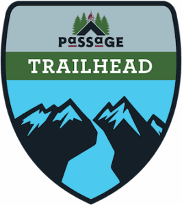 A shield-shaped emblem with mountain silhouettes, the word TRAILHEAD in bold white letters, and the word PASSAGE above, featuring pine trees and a small building at the top.