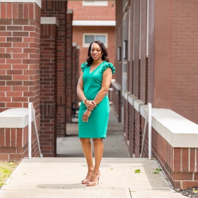 A woman in a green dress and pink heels stands smiling on a walkway between brick buildings, with her hands clasped in front of her.