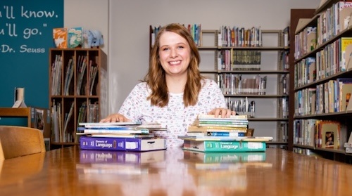 A young woman with long hair smiles while sitting at a library table covered with stacks of books. Shelves of books and a blue wall with a Dr. Seuss quote are visible in the background.
