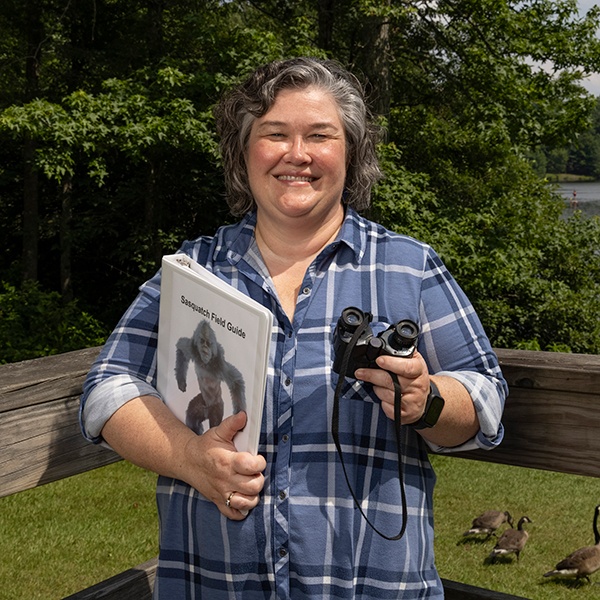 A smiling woman in a blue plaid shirt holds binoculars and a “Sasquatch Field Guide” while standing on a wooden deck outdoors, with trees, grass, and geese in the background.