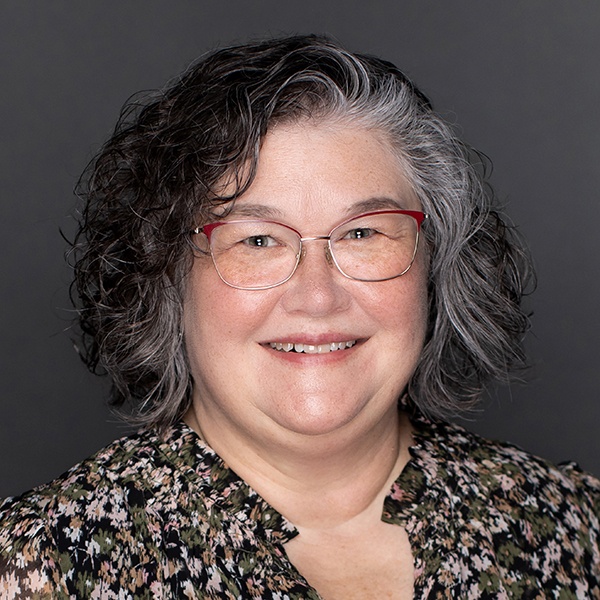 A smiling middle-aged woman with curly gray hair and glasses, wearing a floral blouse, poses in front of a plain dark background.