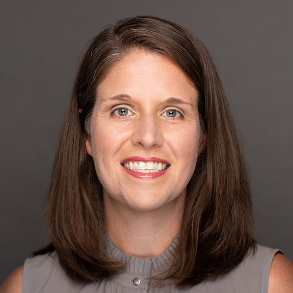 A woman with straight brown hair, wearing a sleeveless gray top, smiles at the camera against a plain dark background.