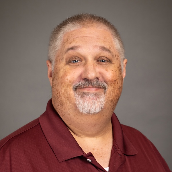 A middle-aged man with short, graying hair and a goatee, wearing a light blue collared shirt, smiles at the camera against a plain gray background.