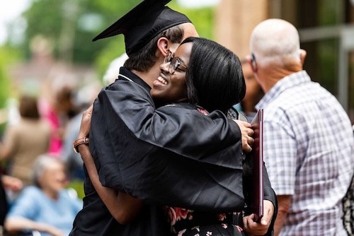 A graduate in a cap and gown hugs a smiling woman outdoors during a graduation ceremony, with other people in the background.