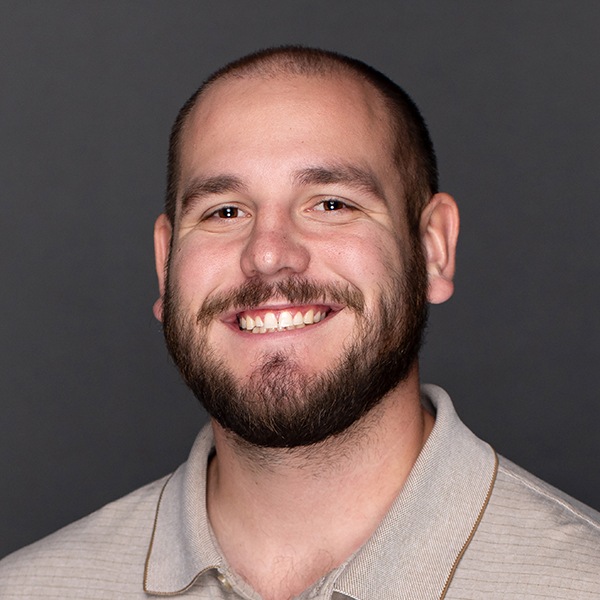 A smiling man with a beard and short hair wearing a light gray collared shirt stands in front of a plain dark gray background.