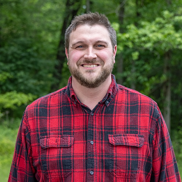 A man with short brown hair and a beard smiles while standing outdoors, wearing a red and black plaid shirt. Green trees and foliage are visible in the background.