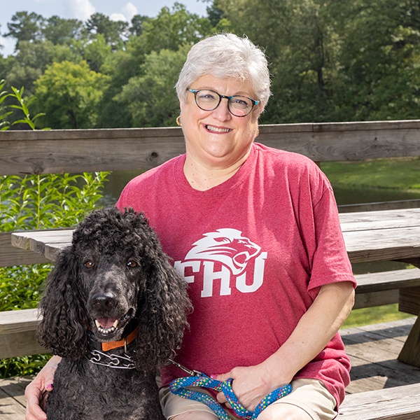 A smiling older woman with short gray hair and glasses sits on a bench outdoors, wearing a red FHU t-shirt and holding a blue leash attached to a black Standard Poodle. Trees and greenery are in the background.