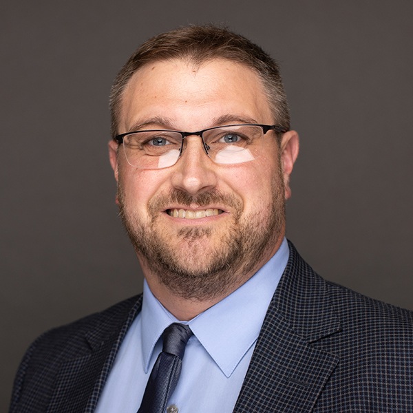 A man with short brown hair, glasses, and a beard, wearing a blue dress shirt, dark patterned suit jacket, and navy tie, smiles in front of a plain gray background.