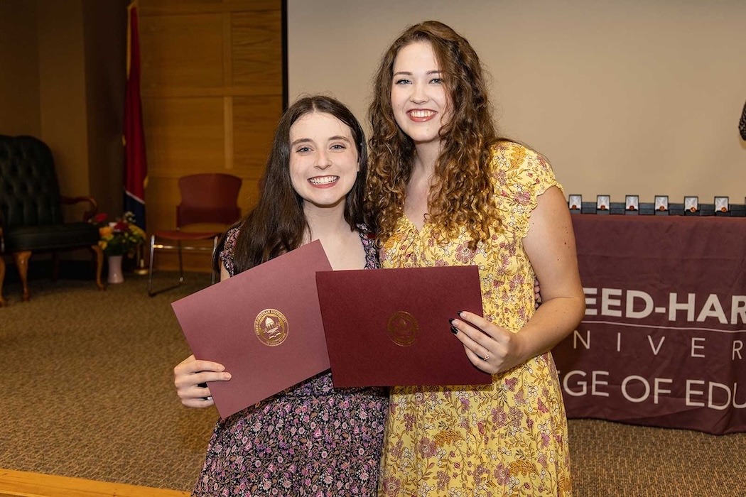 Two young women smiling and holding certificates stand together in a room, likely at a graduation or award ceremony. Both wear floral dresses, and a partial university banner is visible behind them.