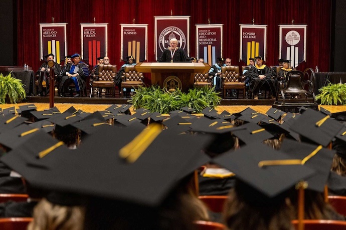 A graduation ceremony with a speaker at a podium on stage, faculty members seated behind, and graduates in caps and gowns facing the stage. Banners with academic departments hang in the background.