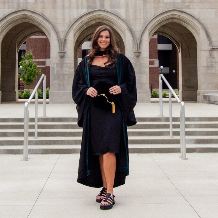 A woman in a black graduation gown and dress stands smiling on steps outside a stone building with arched doorways, holding her graduation cap in front of her.
