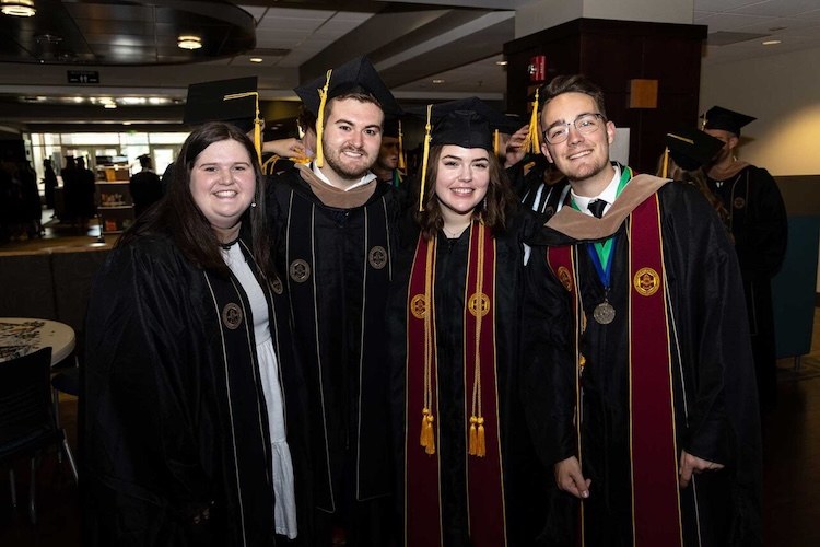 Four graduates wearing black caps and gowns smile and pose together indoors. Two wear maroon stoles and one has medals and cords, indicating academic honors. The setting appears to be a university or college.
